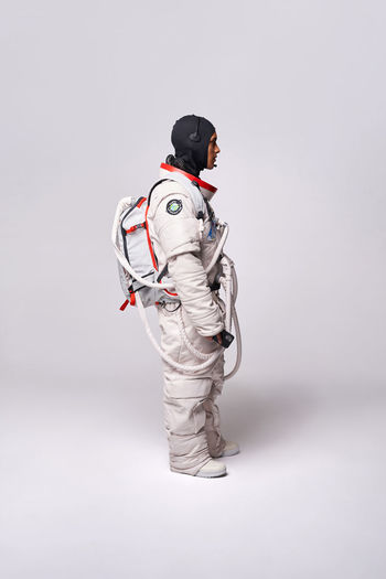 Full body side view of colombian male astronaut in spacesuit with helmet in hand standing in studio against white background and looking away