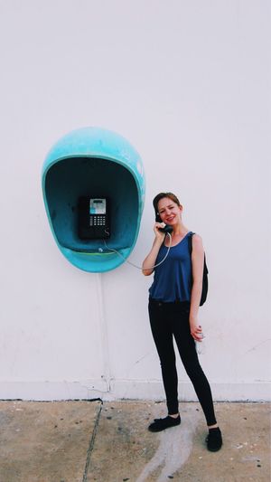 Smiling young woman talking through pay phone while standing against white wall