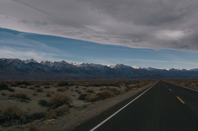 Empty road amidst landscape leading towards mountains against cloudy sky