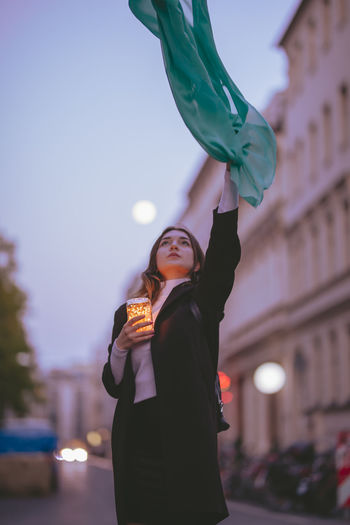 Woman holding illuminated lighting equipment with scarf while standing on road in city during sunset