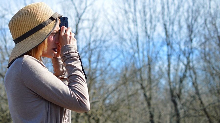 Side view of young woman photographing against bare trees