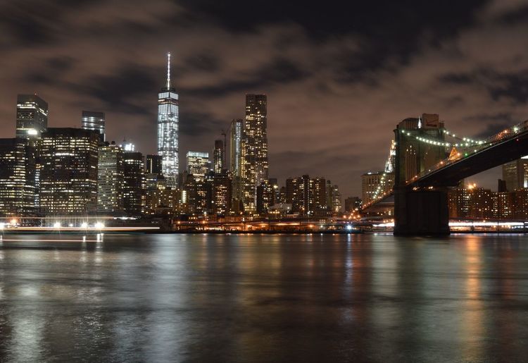 Low angle view of illuminated one world trade center and brooklyn bridge by river