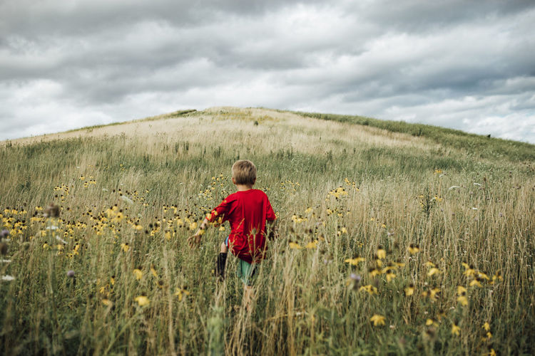 Rear view of boy walking on grassy field against cloudy sky