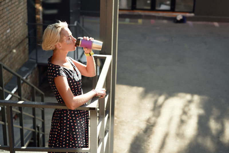 High angle side view of joyful alternative female with dyed hair and in braces standing on stairs with reusable cup of hot takeaway drink and looking at camera