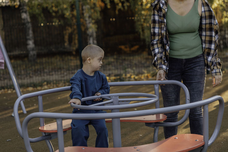 Boy with down syndrome in funny hat walks in the playground with his mother, spinning on a carousel