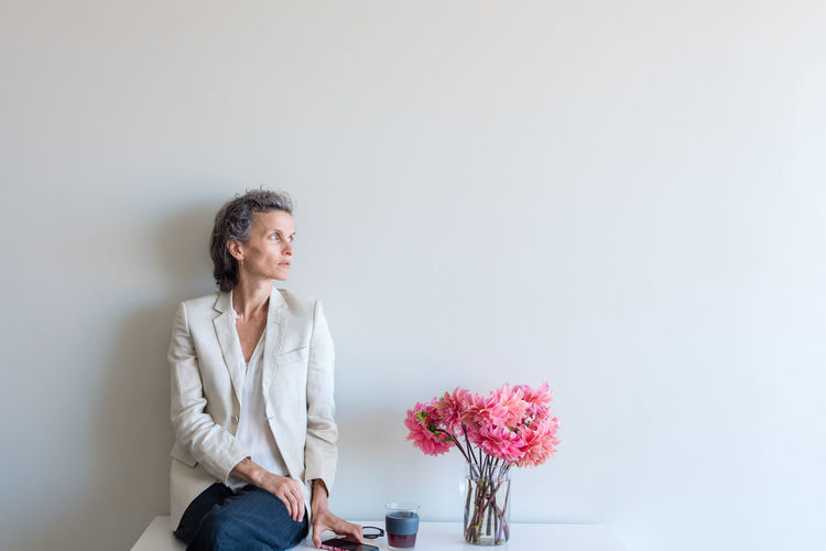 Thoughtful woman sitting by flower vase against wall