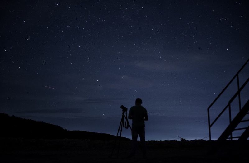 Silhouette man with camera and tripod standing against sky at night
