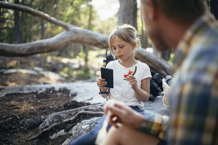 Daughter eating watermelon while using phone by father in forest