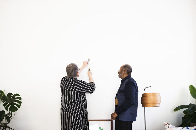 Senior woman hammering nail on wall to fix painting standing by elderly man in living room