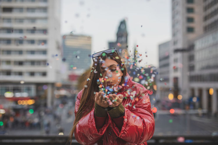 Young woman blowing confetti while standing in city
