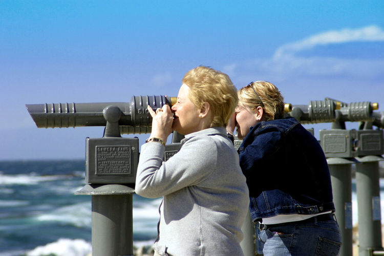 Side view of women using coin-operated binoculars against sky