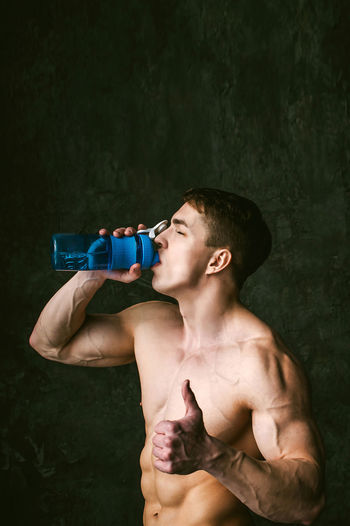 Shirtless man drinking while gesturing thumbs up by wall