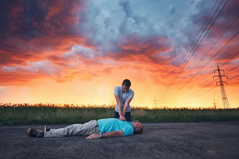 Man resuscitating friend lying on road against cloudy sky during sunset