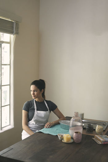 Young woman looking through window while sitting with containers and ingredients at table
