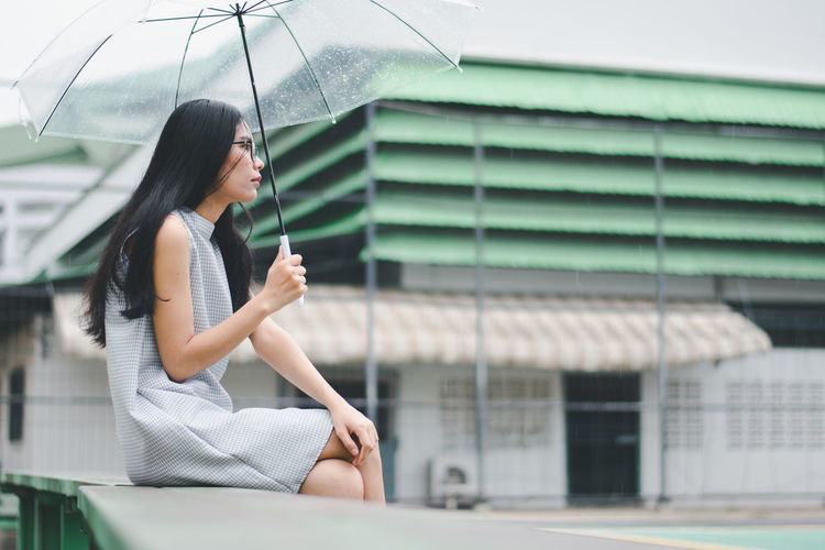 Side view of young woman holding umbrella sitting on retaining wall against building