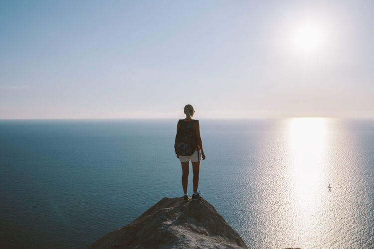 Rear view of young woman standing on cliff by sea against clear sky