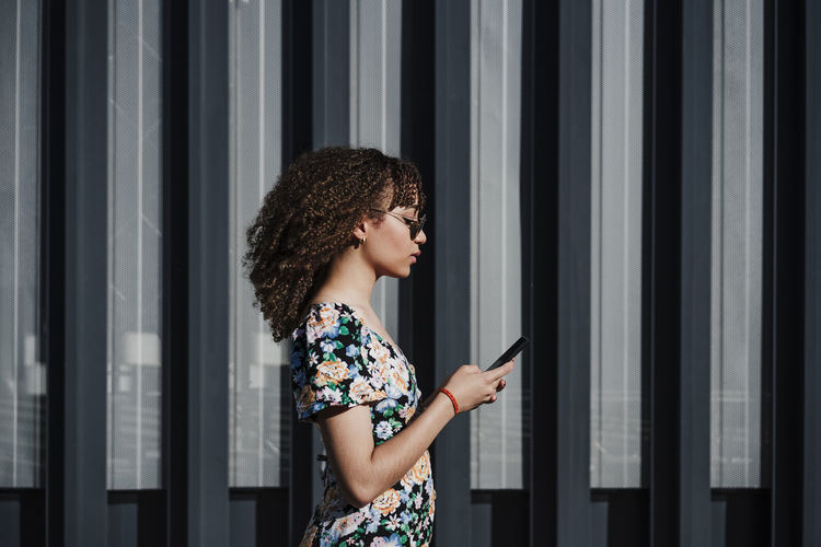 Curly haired woman using smart phone while standing by wall