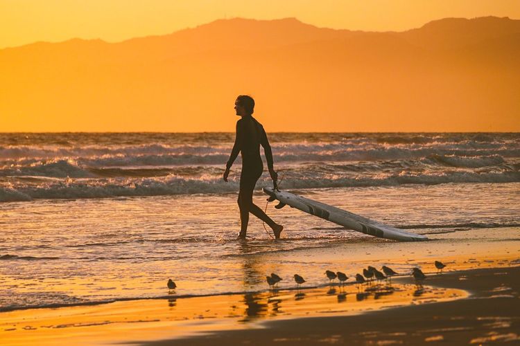 Full length of man with surfboard walking on beach at sunset