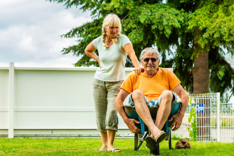 Senior man sitting on wheelbarrow while standing with wife at yard