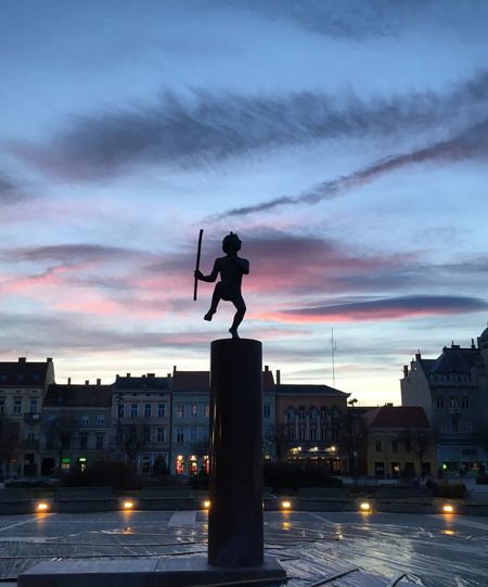 Silhouette statue by illuminated buildings against sky during sunset