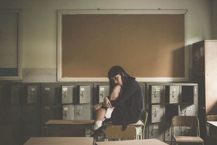 Side view portrait of young woman sitting on desk by lockers
