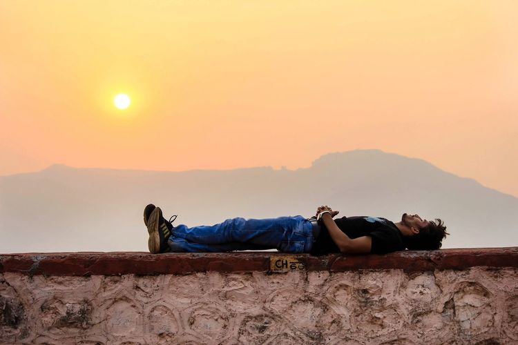 Man sleeping on retaining wall against orange sky