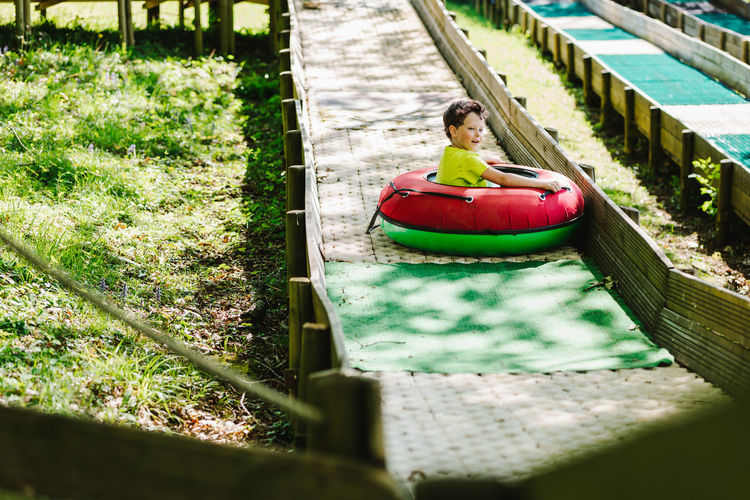 Side view of delighted little child in t shirt smiling while riding inflatable sled tube on slide on playground in sunny adventure park