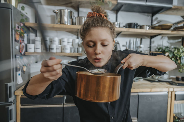 Young female chef tasting broth soup while standing in kitchen