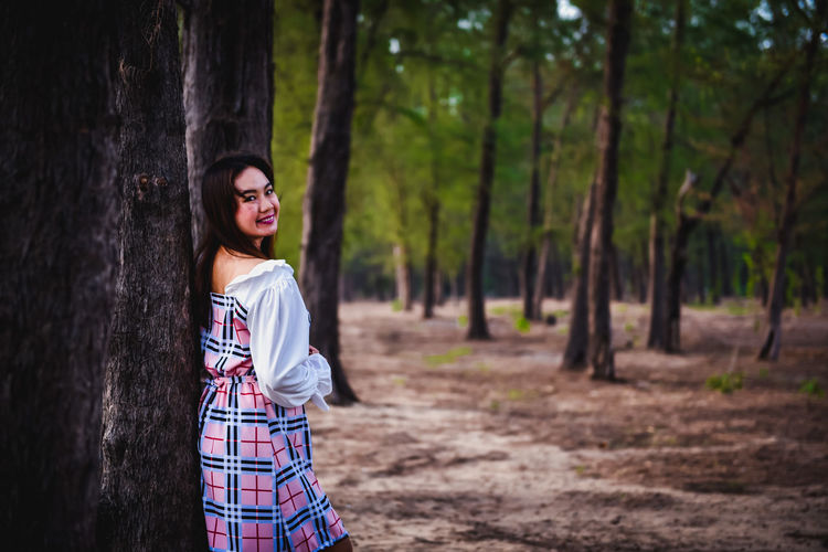 Smiling young woman standing by tree trunk in forest