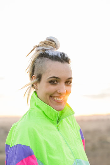 Side view of extraordinary lady with contemporary hairstyle wearing multicolored sportive jacket looking at camera on blurred background of nature