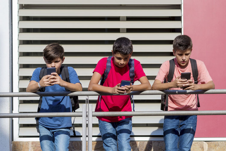 Friends using smart phones while standing by railing in school building during sunny day