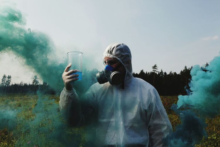 Man looking at liquid in container while wearing gas mask on field against sky