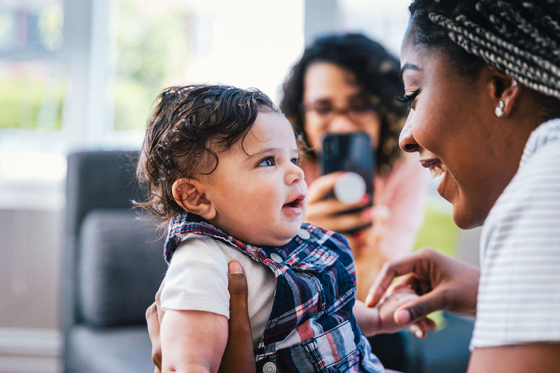 Cheerful mother playing with cute son while girlfriend photographing them in living room