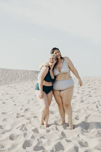 Glad curvy female friends in swimwear stand on sandy seashore and enjoying summer vacation together