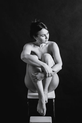 Full length of naked woman sitting on stool against wall