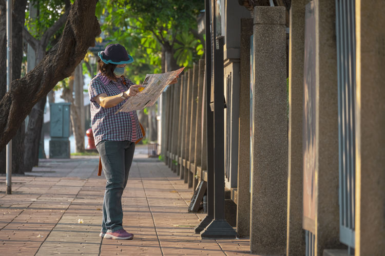 Full length of woman wearing mask reading newspaper while standing on footpath