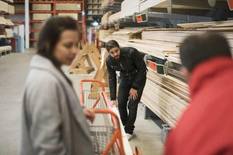 Female customer looking at salesman and man placing wooden plank on trolley in hardware store