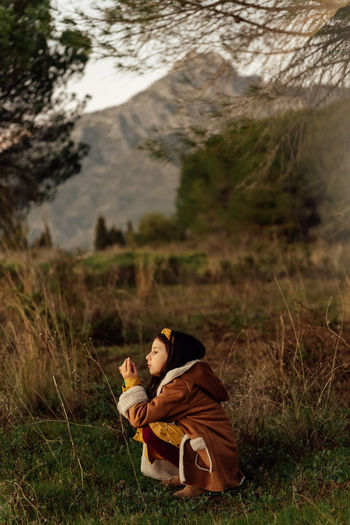 Side view of curious little girl in warm clothes observing environment while spending time alone on grassy meadow against mountains
