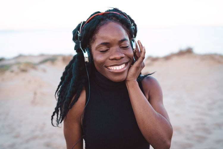 Optimistic african american female in headphones enjoying songs and dancing with closed eyes on sandy shore against sea in summer evening