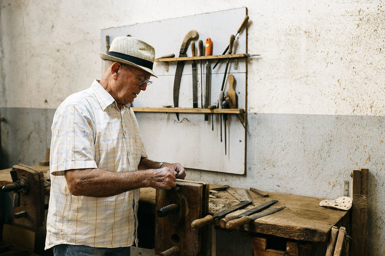 Side view of senior male carpenter grinding wooden details while making handmade folding fans in messy workshop