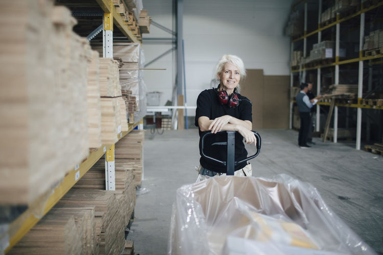 Portrait of smiling worker leaning on pallet jack by wooden planks at industry
