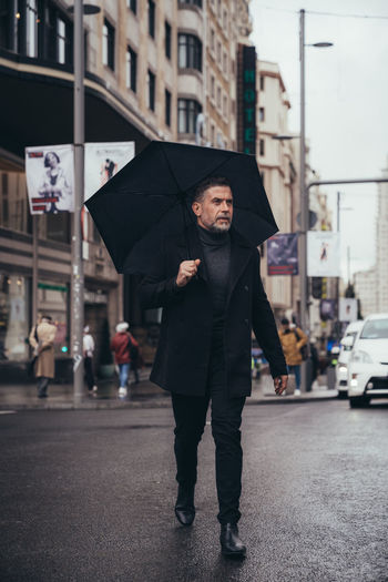 Pensive successful mature bearded male entrepreneur in black standing under umbrella during walk in street in rainy day