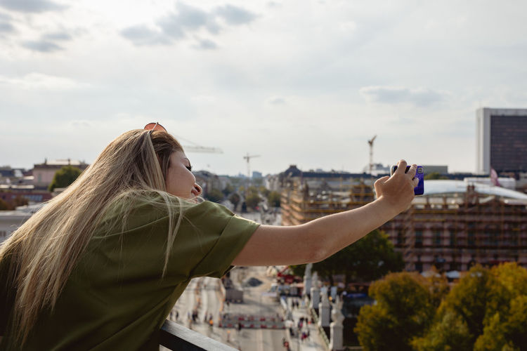 Young woman photographing in city against sky