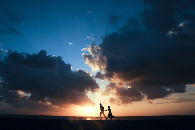 Silhouette couple running at beach against cloudy sky