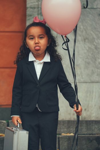 Portrait of smiling girl holding balloon and briefcase while sticking out tongue