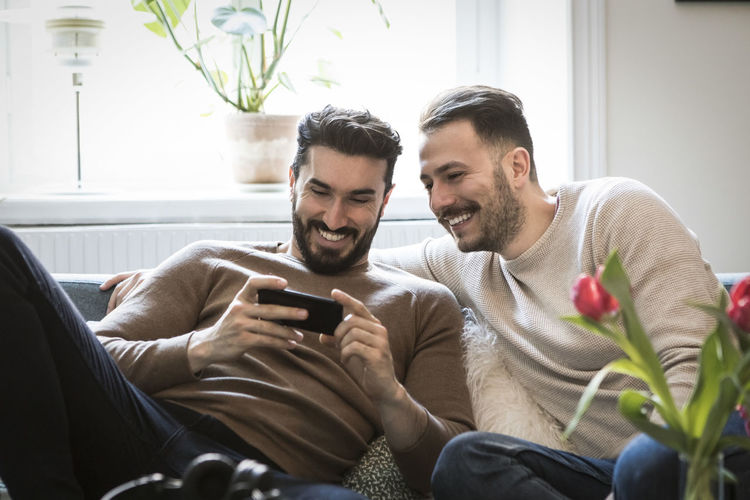 Smiling young men looking at smart phone while sitting on sofa at home