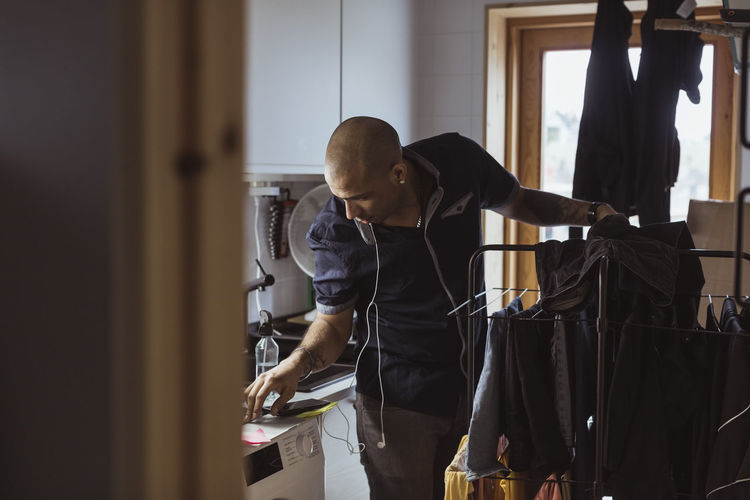 Bald man looking at washing machine while doing laundry chores in utility room