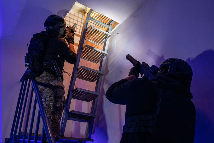 Back view of unrecognizable male soldiers in protective gear and helmet standing on staircase and taking aim with rifles