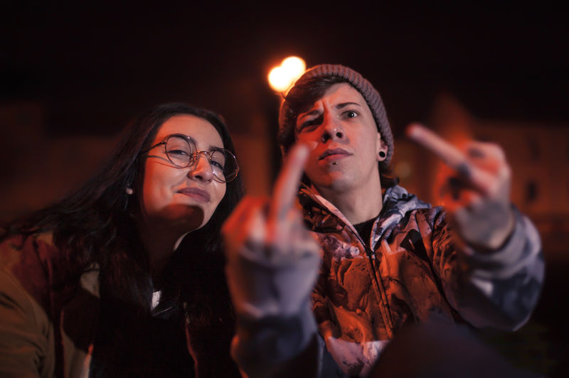 Portrait of young man showing middle fingers by friend in illuminated city at night