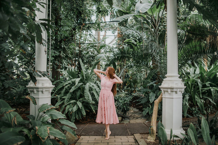 Full body of redhead female standing on tiptoes with hands on head among tropical palm and plants in glasshouse